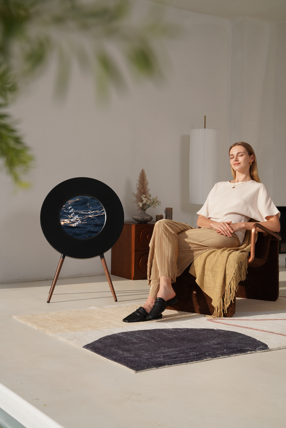A woman sits in a modern living room setting next to a black circular speaker on a tripod stand, which features a circular ocean scene printed in the center.