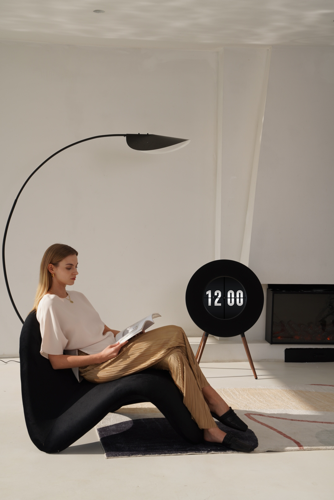 A woman sits in a black sculptural lounge chair reading a magazine in a minimalist, sunlit room. A round black digital clock on wooden legs stands nearby, and a large black floor lamp curves over her.