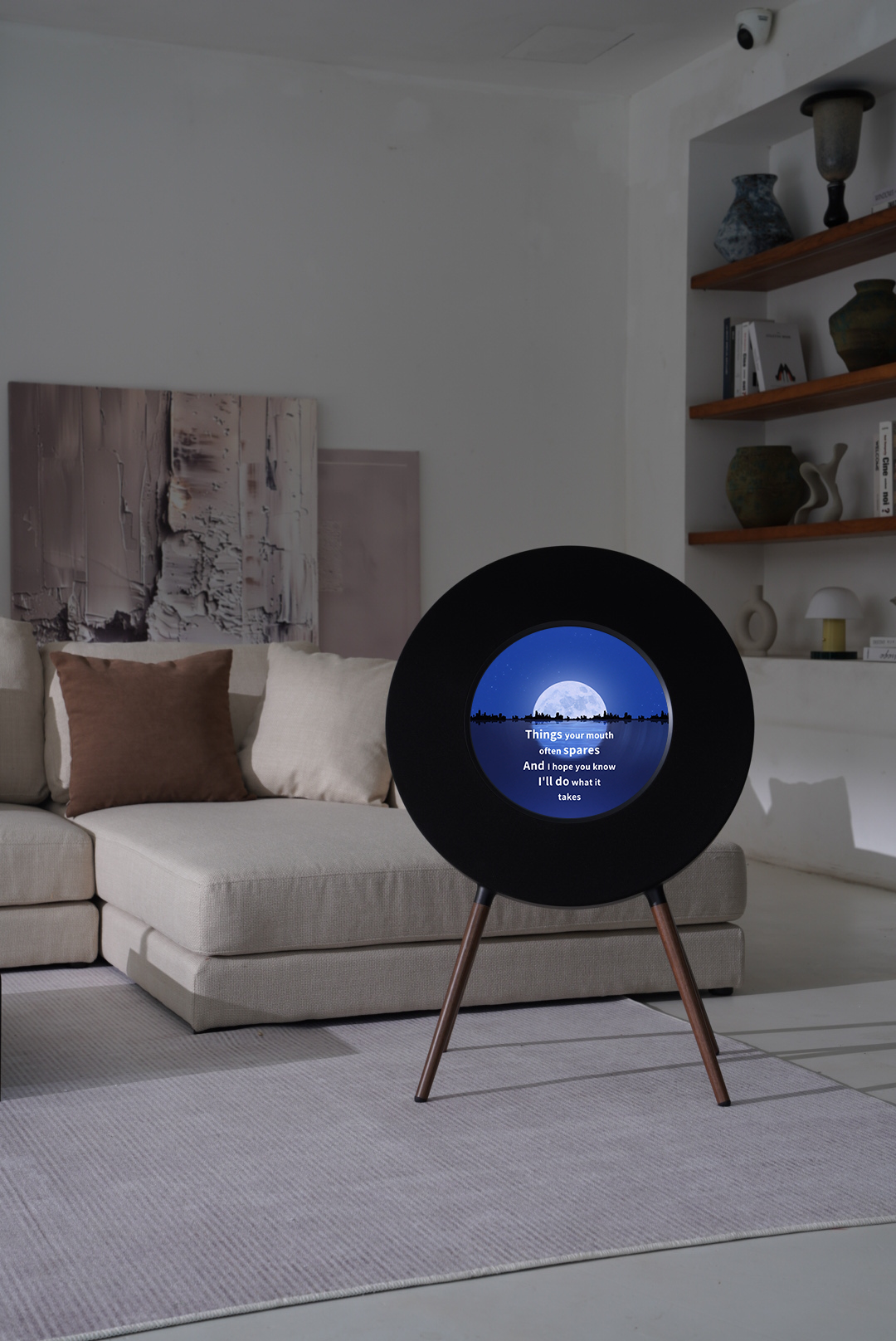 A black circular speaker on a tripod stand in a modern living room, displaying a blue and white moonlit sky design on its front screen.